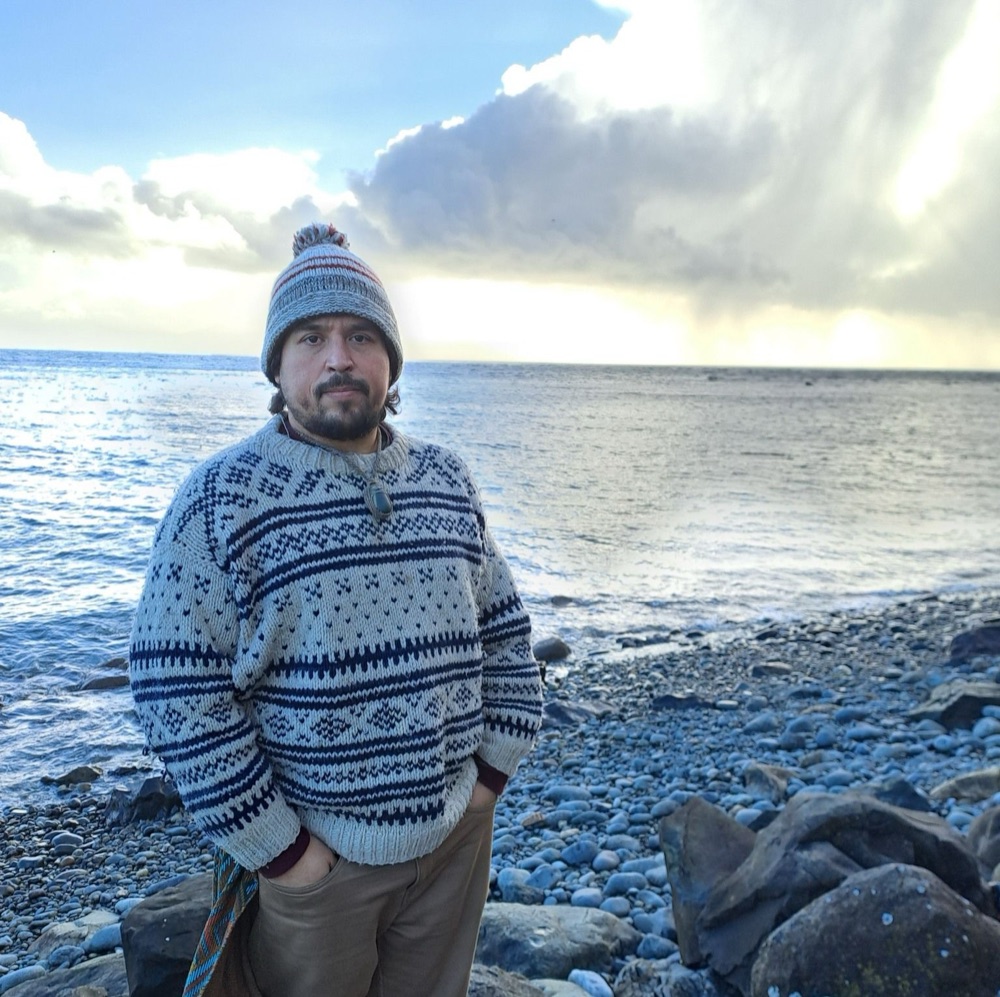 Jorge Pedret standing on a pebble beach in a knitted hat and sweater, with the sea and a storm cloud behind him.